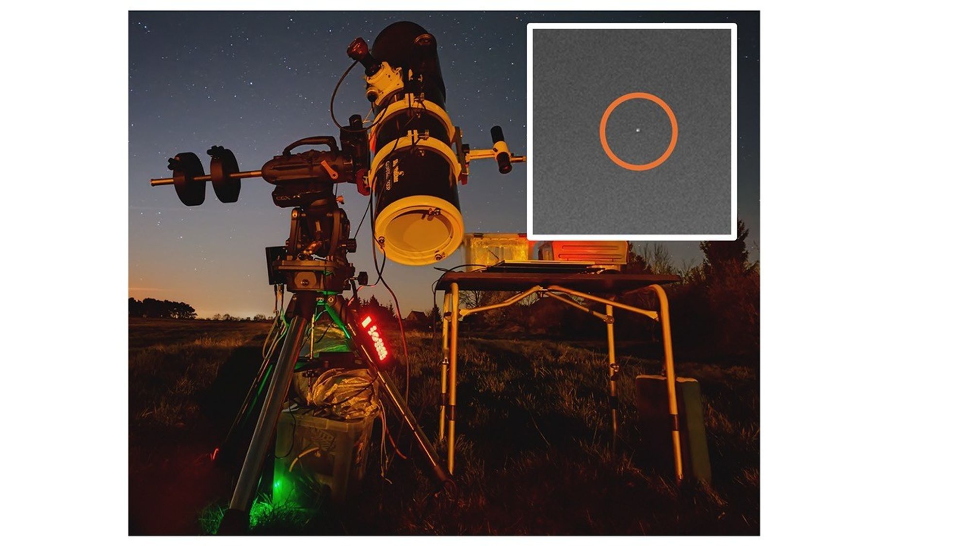 A telescope mounted on a tripod stands outdoors at night under a starry sky, illuminated by warm orange and red lights. Nearby, a small table holds equipment. In the top-right corner, an inset zoomed image shows a faint point of light circled in orange against a grainy background, highlighting a small source of light on the Moon’s surface.